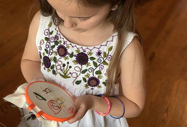A young girl in a white embroidered dress practicing simple embroidery on a piece of cloth held in a hoop