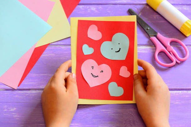 A child's hands holding a handmade greeting card with red and light blue paper hearts and smiling faces