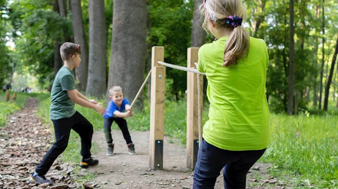 Three children playing tug-of-war with a thick rope outdoors in a sunny, green forest or park